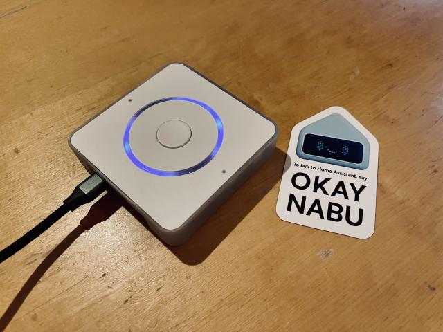 A white square object sitting on a wooden table, next to a cute sticker that says Okay Nabu.